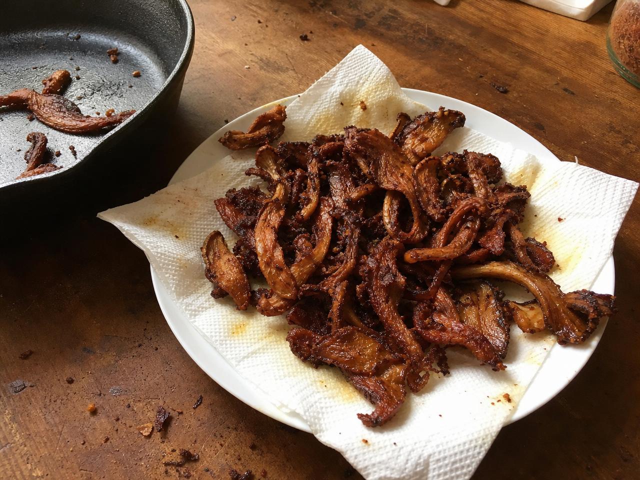 A plate of crispy, cooked mushroom strips on a paper towel next to a cast iron skillet.