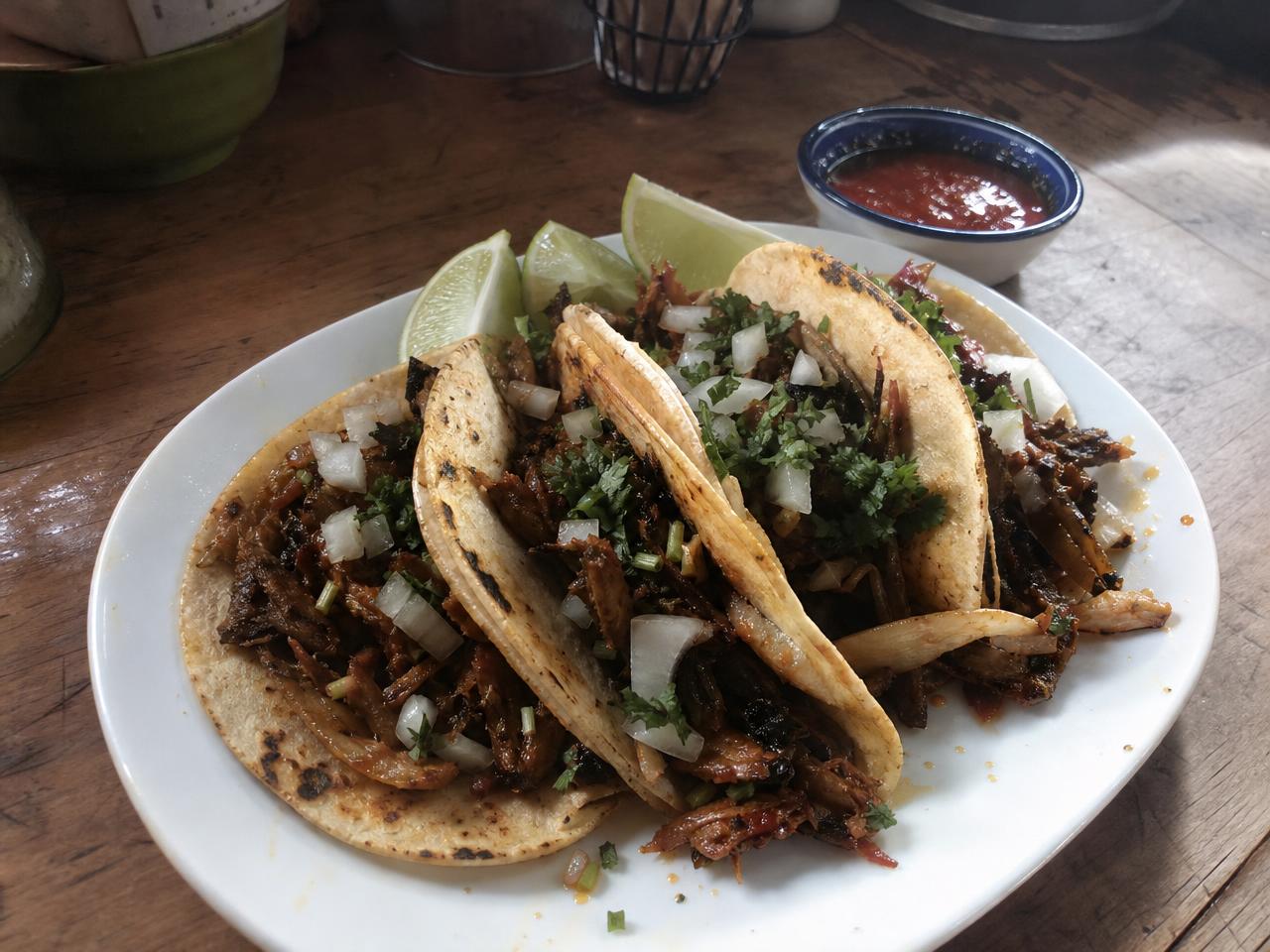 Three tacos on a white plate with lime wedges and a small bowl of salsa in the background.